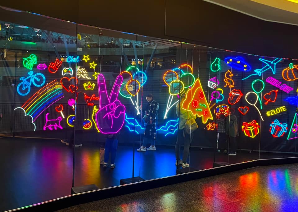 Neonowa instalacja artystyczna z motywami tęczy, roweru, pizzy i symbolu pokoju za szkłem, odbijająca sylwetki przechodniów.
