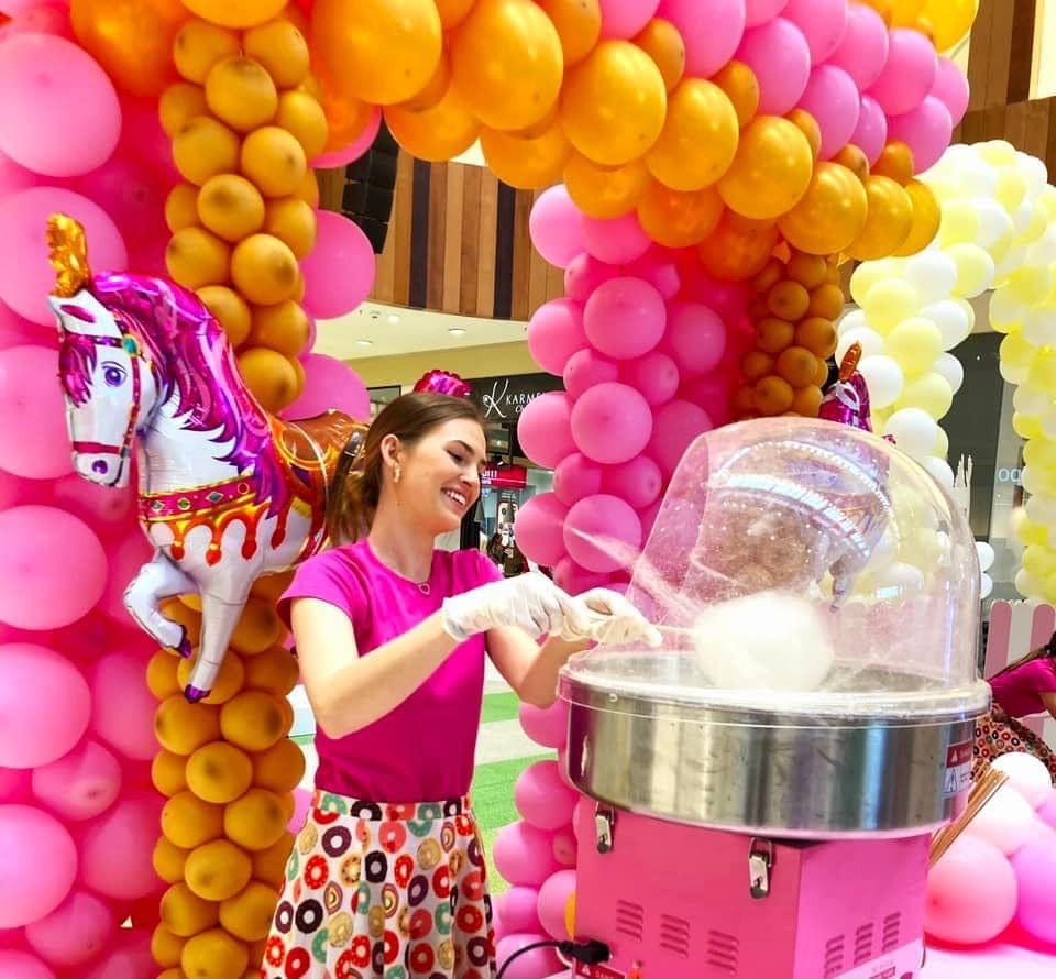 Uśmiechnięta kobieta w rękawiczkach przygotowuje watę cukrową na różowym wózku, otoczona dekoracjami z różowych i żółtych balonów oraz balonem w kształcie konia, w tle widoczny fragment galerii...