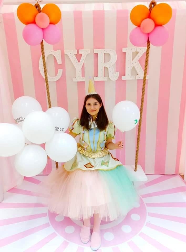 Uśmiechnięta kobieta w kostiumie cyrkowym siedzi na huśtawce z białymi balonami, na tle różowo-białego pasiastego tła z napisem 'CYRK' i balonami pomarańczowo-różowymi.