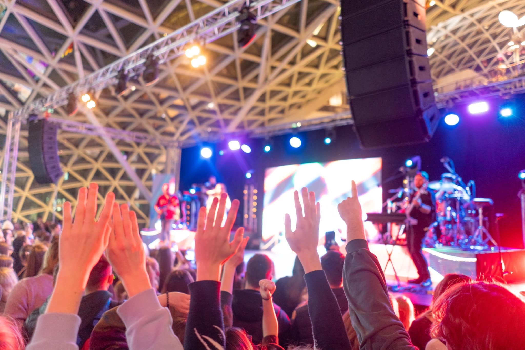 Koncert z widokiem na scenę z podniesionymi rękami publiczności, oświetloną przez reflektory i dekoracyjną konstrukcją sufitową w tle.