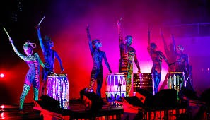 Grupa tancerzy z pomalowanymi ciałami i podświetlonymi bębnami na scenie w intensywnym, kolorowym oświetleniu.