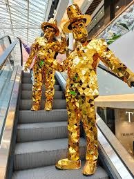 Dwie osoby w całości ubrane w błyszczące, złote kostiumy z geometrycznych elementów, stojące na ruchomych schodach w centrum handlowym. Jeden z performerów unosi rękę w geście powitania.