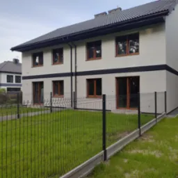 Nowoczesny, dwukondygnacyjny dom z szarą elewacją i czarnym pasem, otoczony metalowym ogrodzeniem panelowym z betonową podmur&oacute;wką, widok z zewnątrz na trawnik.