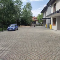 Ułożona kostka brukowa na podjeździe do nowych domów szeregowych, z zaparkowanym niebieskim samochodem osobowym i widocznymi skrzynkami gazowymi w kolorze żółtym.