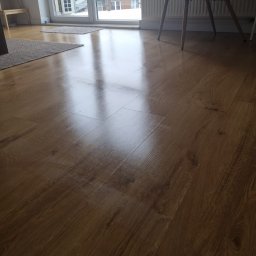 Asia Asia - Błyszcząca, świeżo umyta podłoga z paneli drewnopodobnych, odbijająca światło z okna balkonowego, z widocznymi dywanikami i fragmentem krzesła.