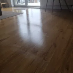 Błyszcząca, świeżo umyta podłoga z paneli drewnopodobnych, odbijająca światło z okna balkonowego, z widocznymi dywanikami i fragmentem krzesła.