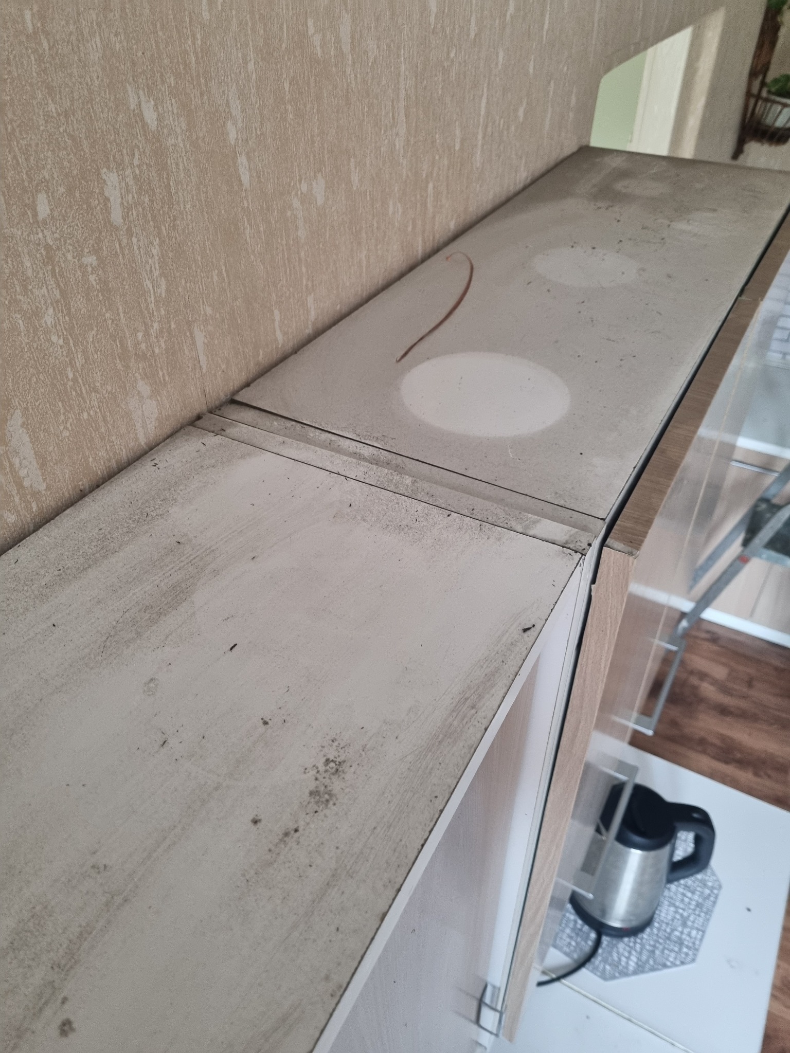 Zabrudzona, zakurzona powierzchnia szafki kuchennej z widocznymi plamami, kurzem i resztkami, z czajnikiem elektrycznym w tle.