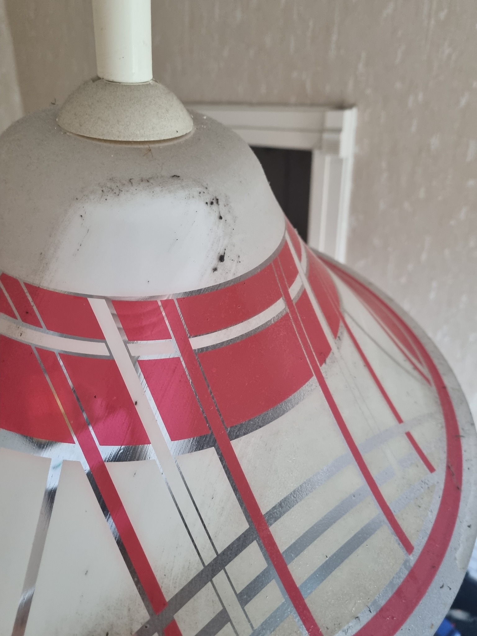 Bliski plan zakurzonego klosza lampy z geometrycznym wzorem w kolorze czerwonym i srebrnym, przed ścianą w neutralnym odcieniu.