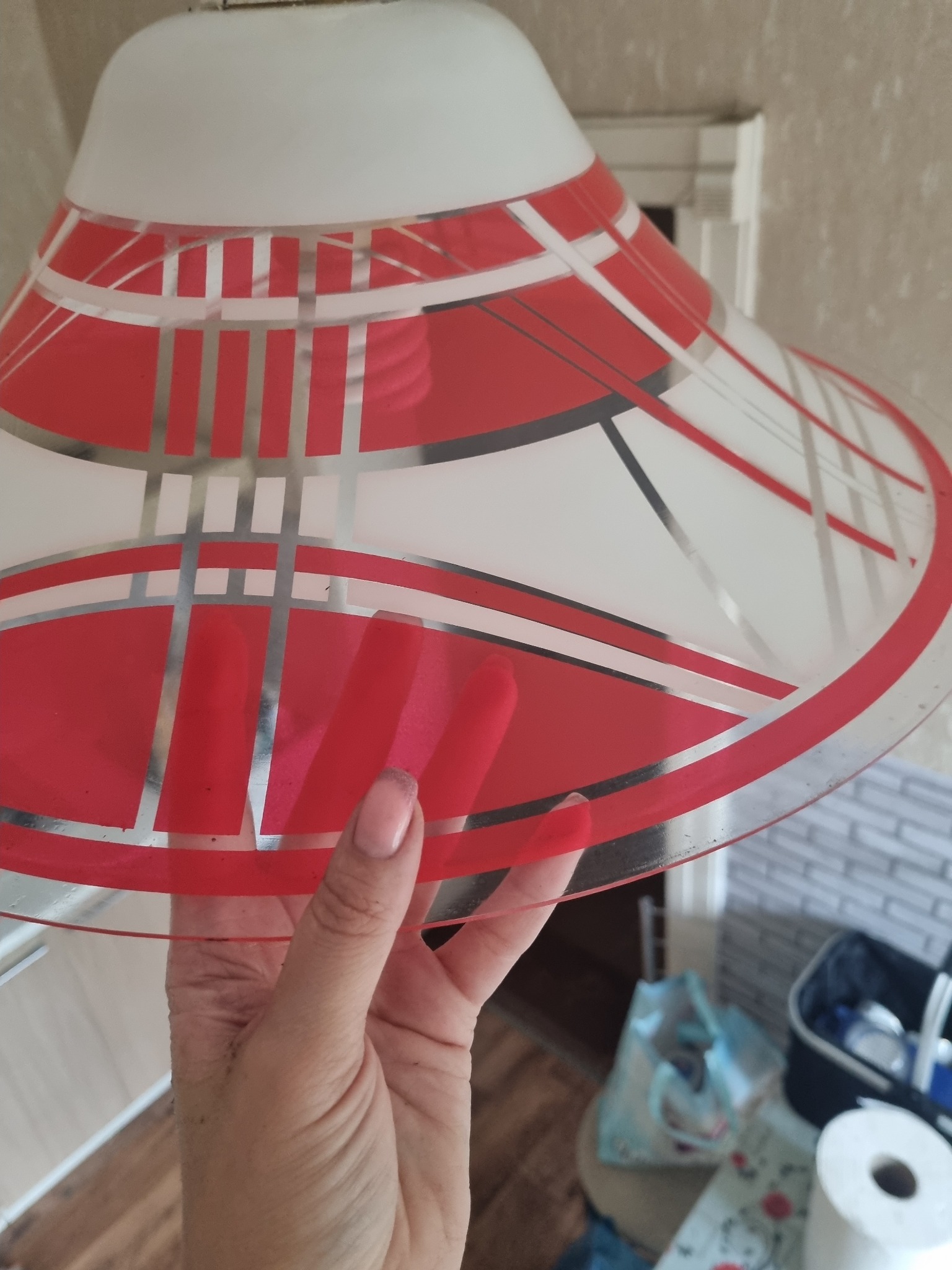 Szklany, biało-czerwony klosz lampy z geometrycznym wzorem, trzymany w dłoni, widoczne zabrudzenia na kloszu i w tle.