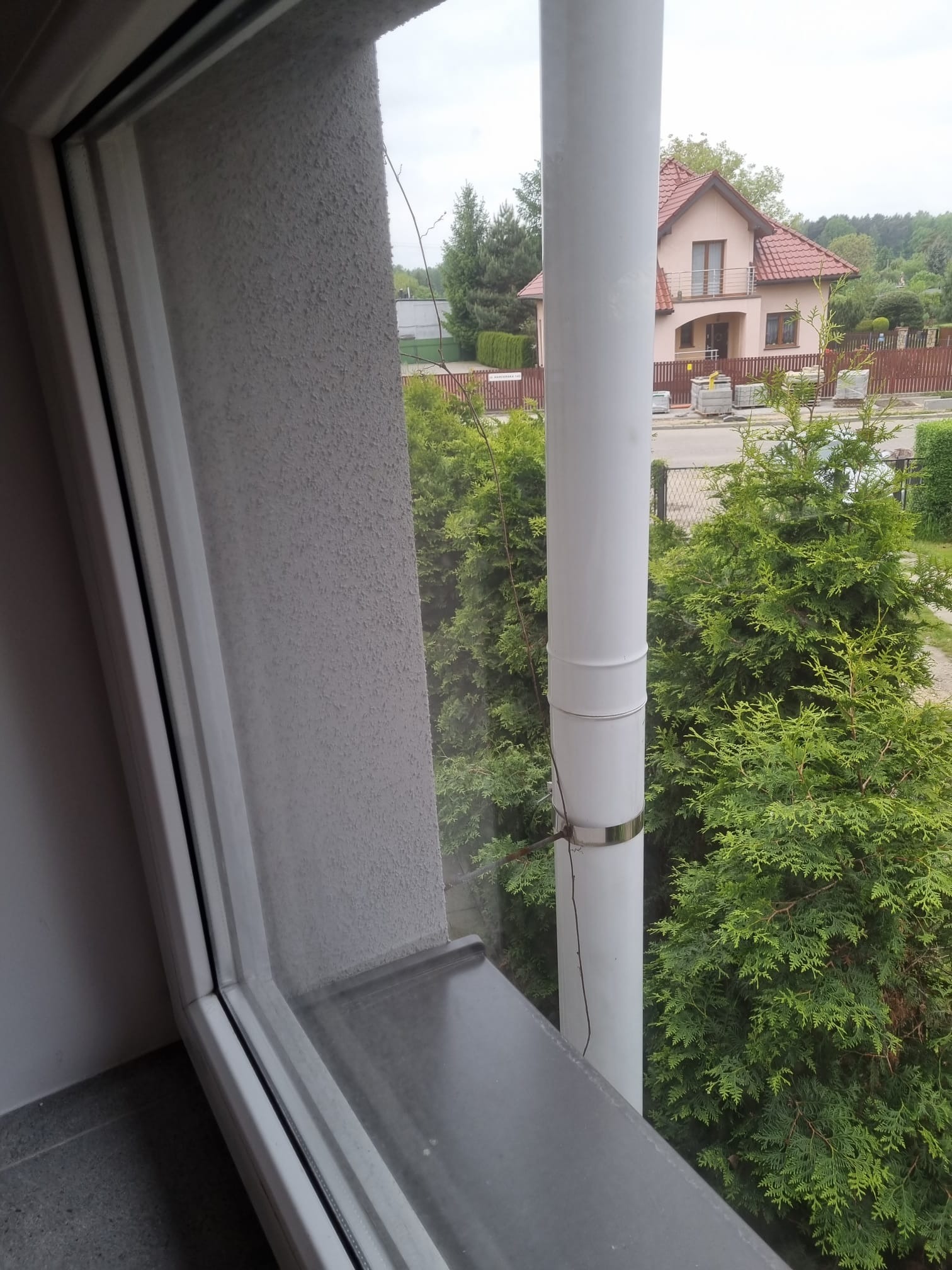 Widok z okna na zewnątrz z widocznym fragmentem rynny spustowej, iglakami i domem w tle.