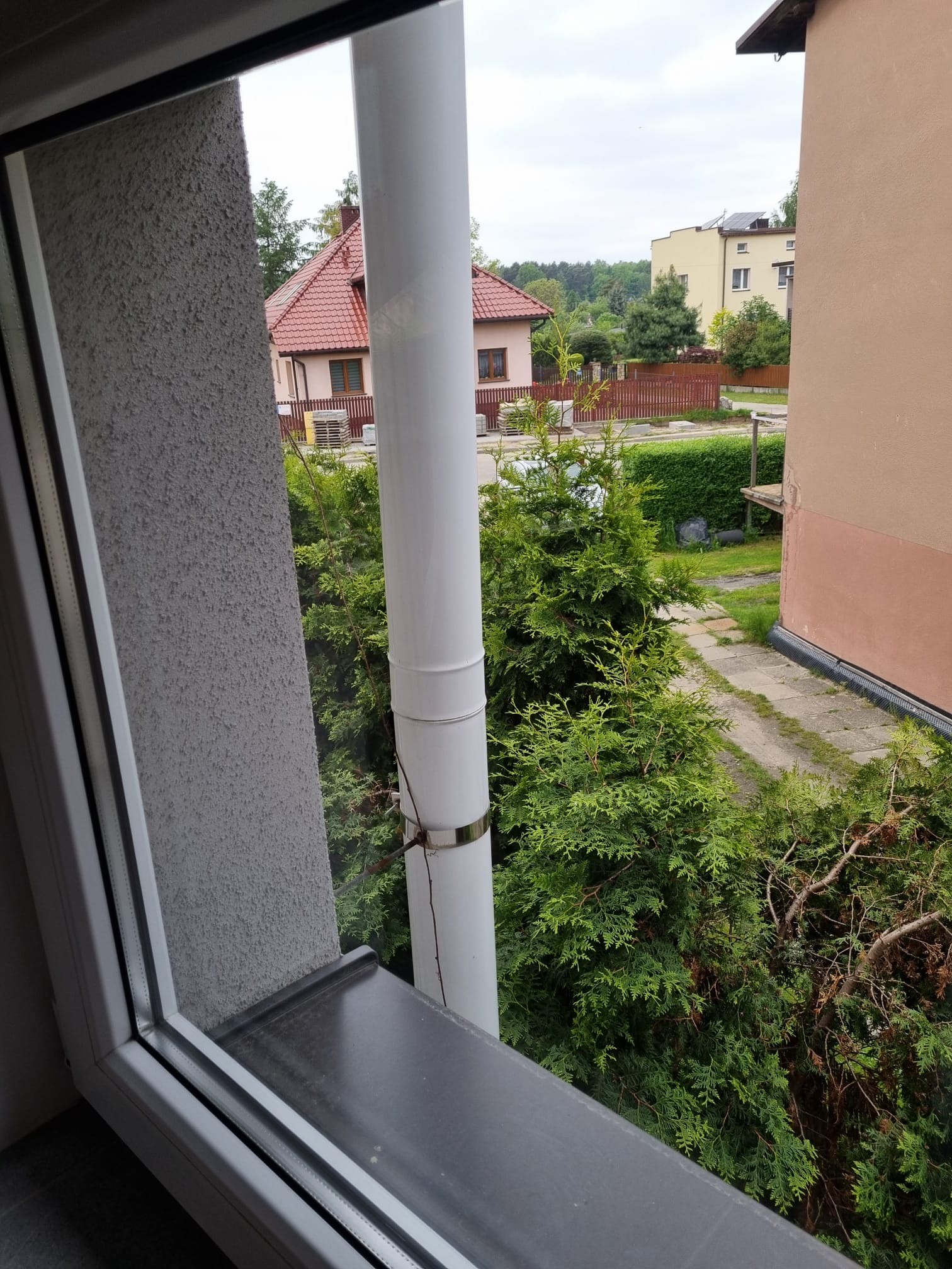 Widok z okna na ogród z iglakami, rynna na pierwszym planie, w tle zabudowa jednorodzinna i wielorodzinna.