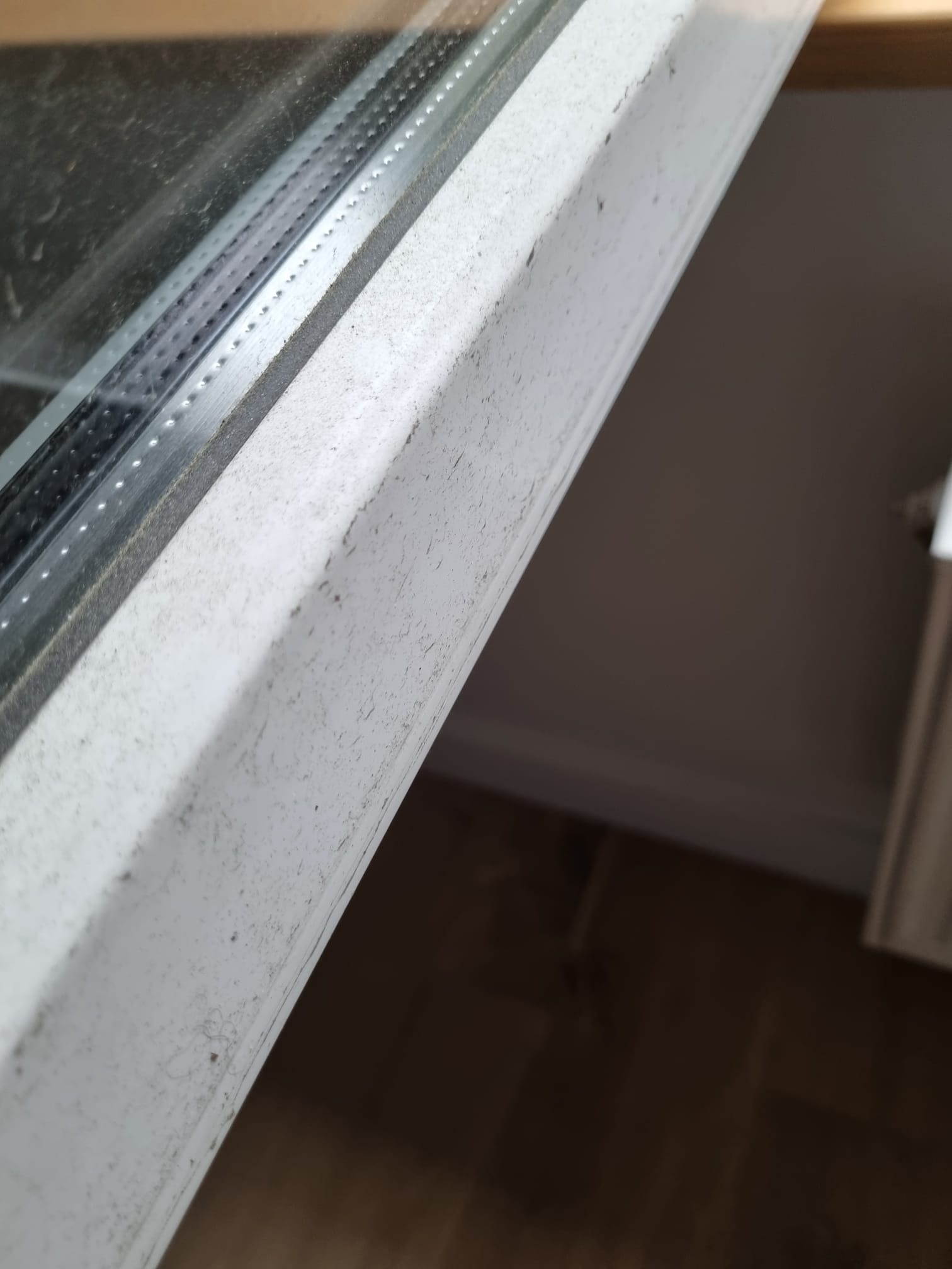 Brudna, biała rama okienna z widocznymi smugami i kurzem, z fragmentem szyby i parapetu oraz podłogą w tle.