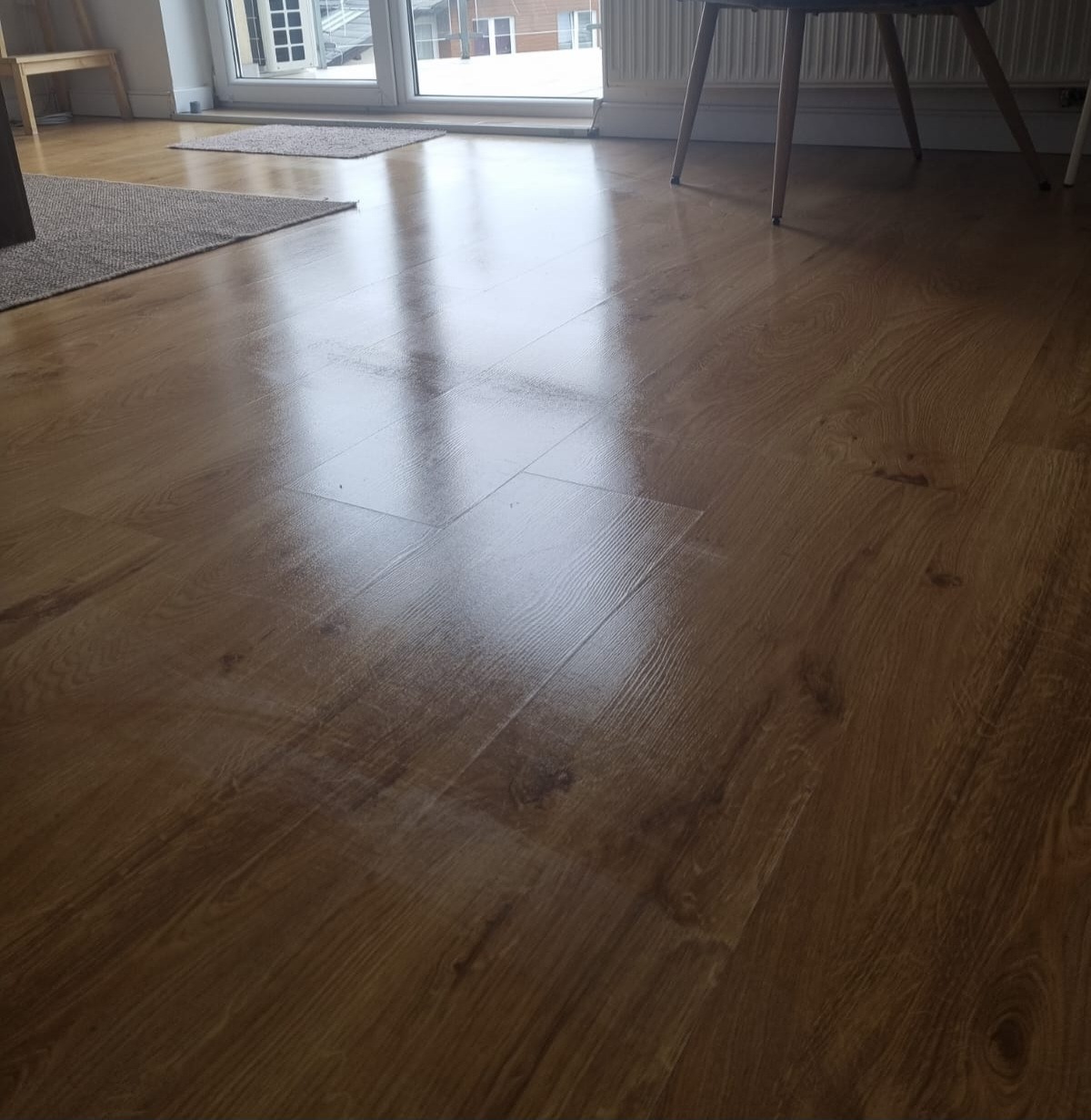 Błyszcząca, świeżo umyta podłoga z paneli drewnopodobnych, odbijająca światło z okna balkonowego, z widocznymi dywanikami i fragmentem krzesła.