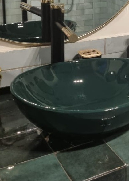 Zielona, owalna umywalka nablatowa z czarną baterią z elementami w kolorze złota, zamontowana na blacie z zielonych płytek.