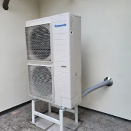 Biała, dwuczęściowa jednostka zewnętrzna z logo Panasonic, zamontowana na białym metalowym stelażu przy jasnej ścianie z widocznymi rurami wentylacyjnymi i odpływowymi.