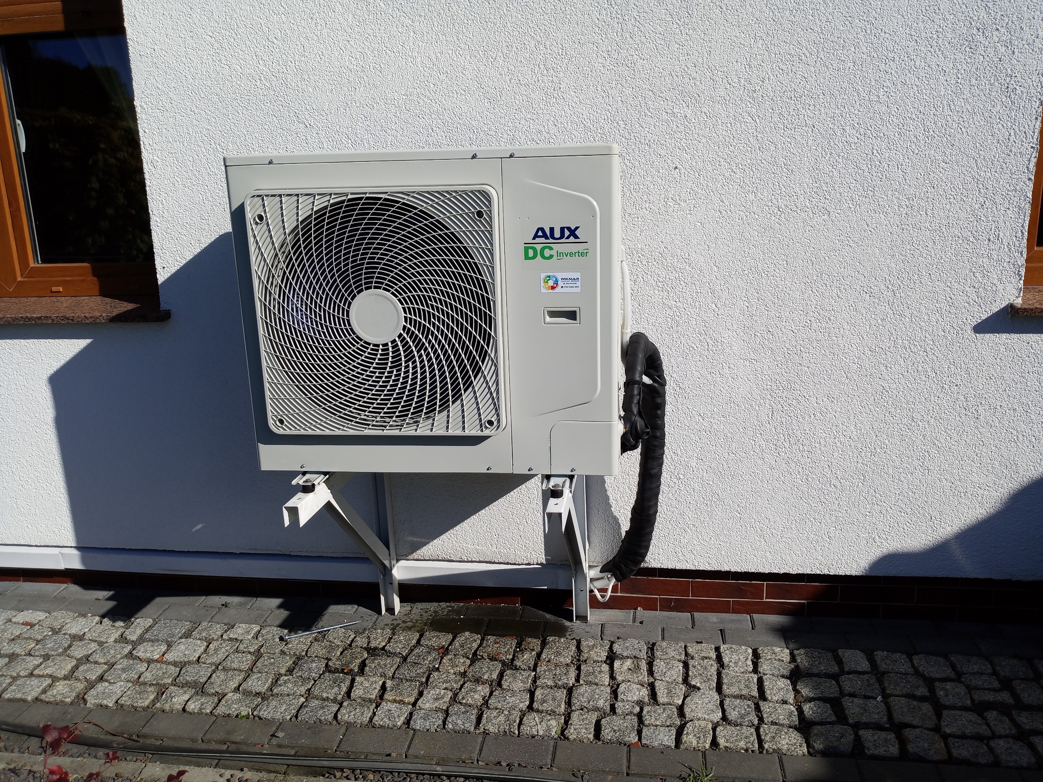 Zewnętrzna jednostka klimatyzacji AUX DC Inverter zamontowana na białej, teksturowanej ścianie budynku. Widoczna kostka brukowa i fragment okna.
