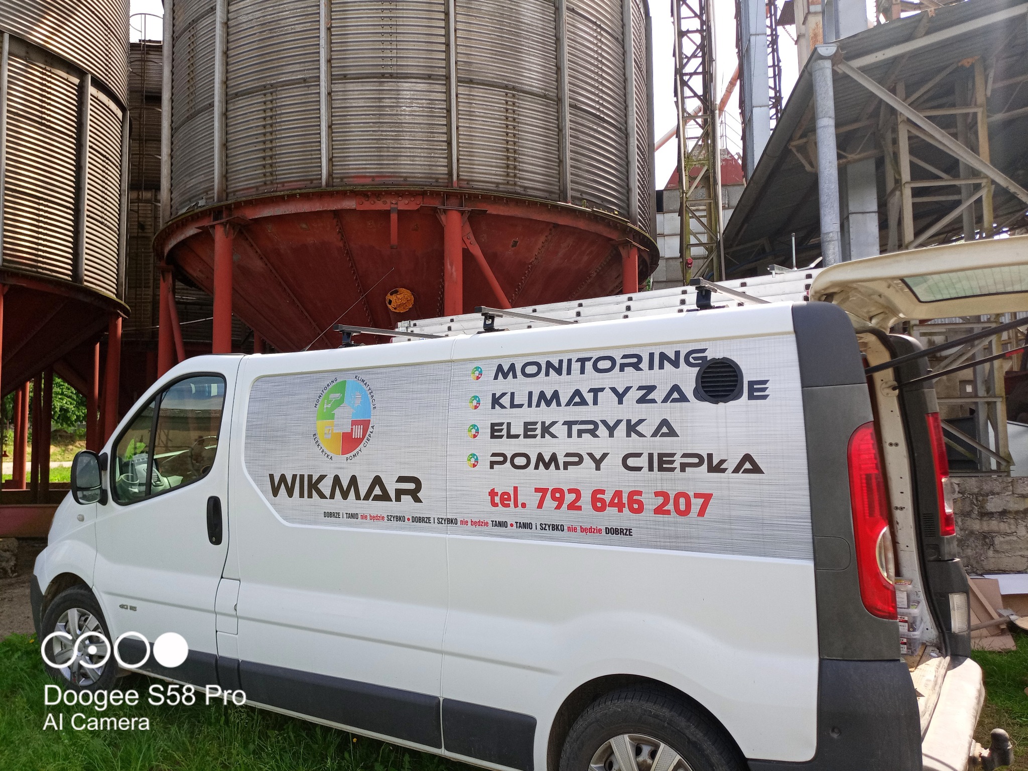 Biały samochód dostawczy firmy Wikmar z reklamą usług: monitoring, klimatyzacja, elektryka, pompy ciepła, zaparkowany na tle silosów zbożowych.