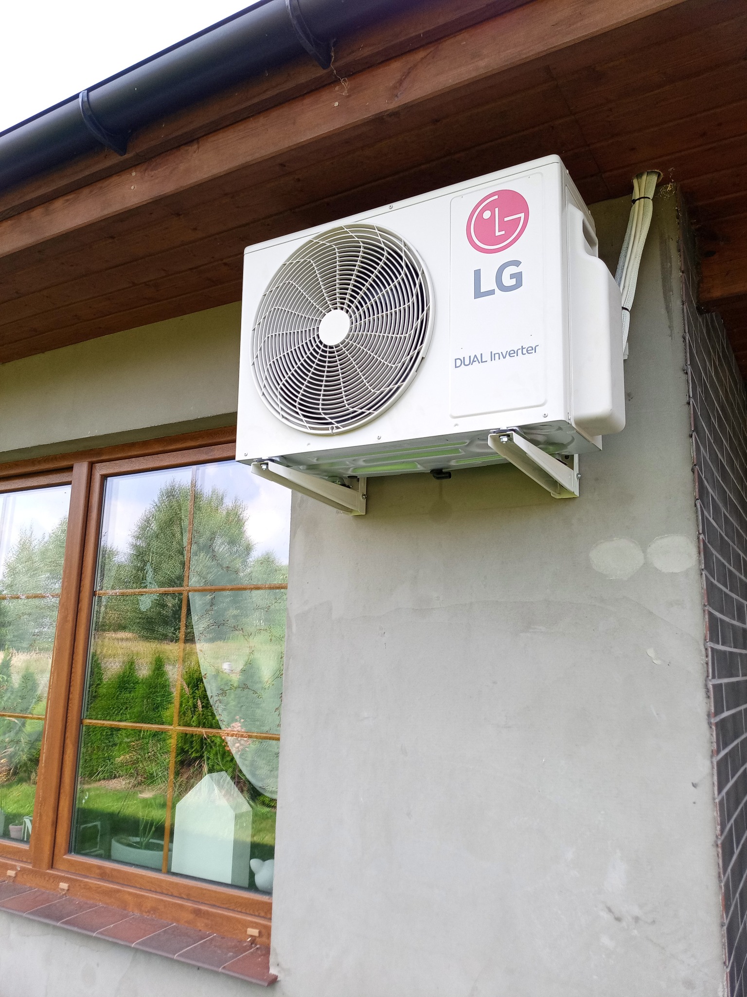 Zewnętrzna jednostka klimatyzacji LG Dual Inverter zamontowana na ścianie budynku, widok z dołu, obok okna z odbijającym się ogrodem.