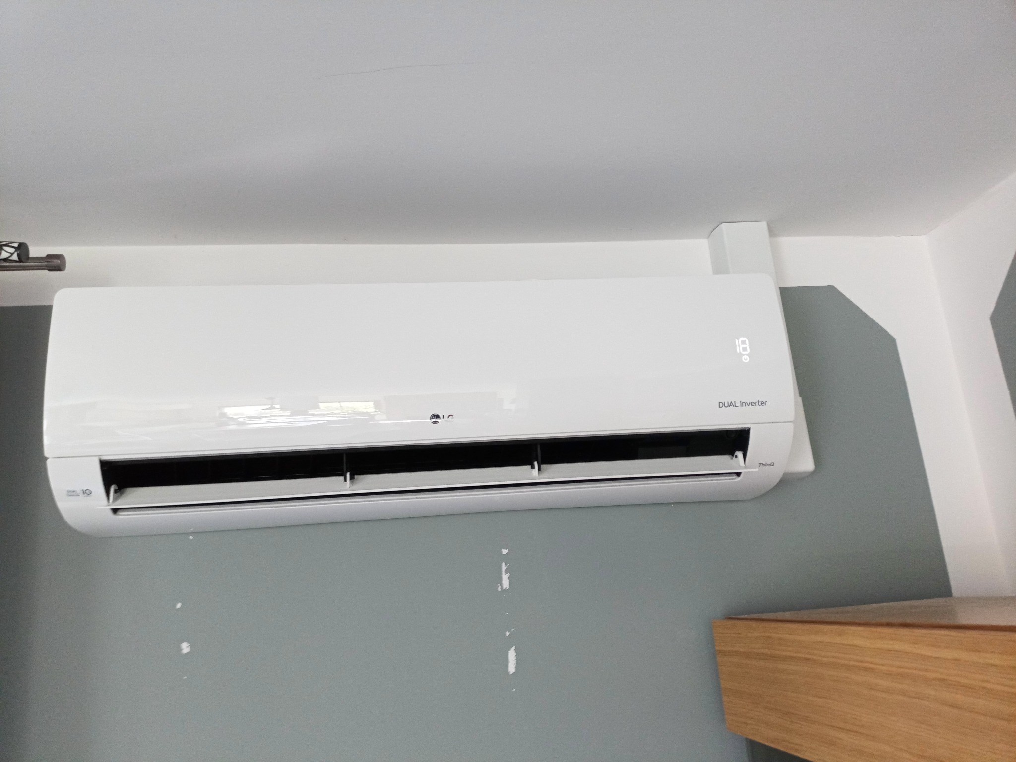 Biała jednostka wewnętrzna klimatyzacji LG Dual Inverter zamontowana na szaro-białej ścianie, z widocznym wyświetlaczem temperatury ustawionym na 18 stopni Celsjusza, z bliska widoczne...