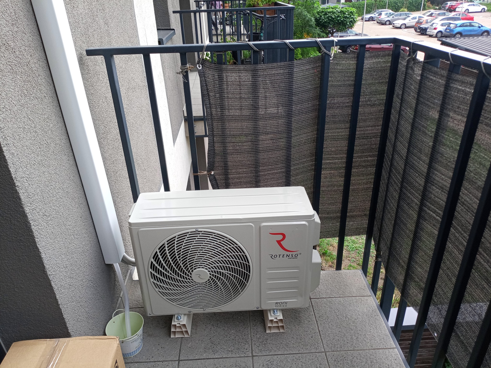 Jednostka zewnętrzna klimatyzacji Rotenso, model Roni Series, zamontowana na balkonie, z odprowadzeniem skroplin do ozdobnego wiadra, widoczna barierka i przesłona balkonowa.