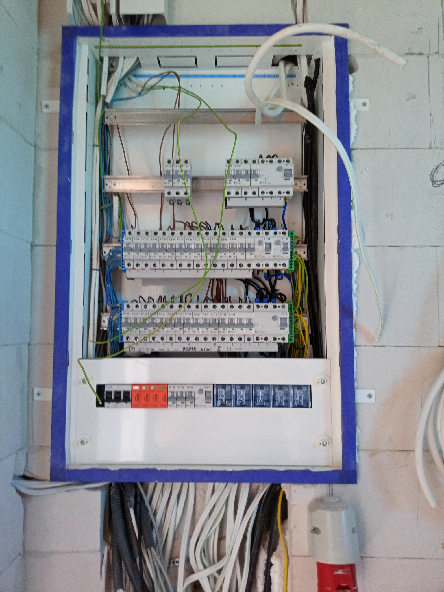 Rozdzielnica elektryczna w trakcie montażu w surowym wnętrzu, widoczne liczne przewody i zabezpieczenia, niebieska obudowa rozdzielnicy, ściana z białych bloczków.