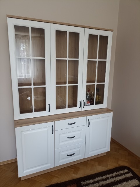 Biały kredens z przeszklonymi drzwiczkami i drewnianym blatem, trzy szuflady i szafki dolne, czarne uchwyty, w tle widoczne butelki.