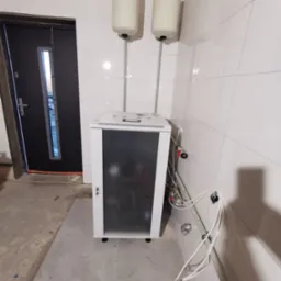 Biała obudowa urządzenia grzewczego z czarnym panelem, dwa białe zbiorniki ciśnieniowe z manometrami na górze, białe płytki na ścianach, surowy beton na podłodze, widoczne rury i zawory.