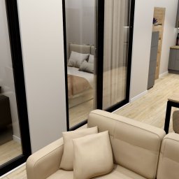 Projekt Kwadrat Studio Aranżacji Wnętrz - Wnętrze mieszkania z widokiem na sypialnię przez szklane drzwi, beżowa sofa z poduszkami na pierwszym planie, w tle kuchnia z szarymi szafkami i drewnianym blatem.