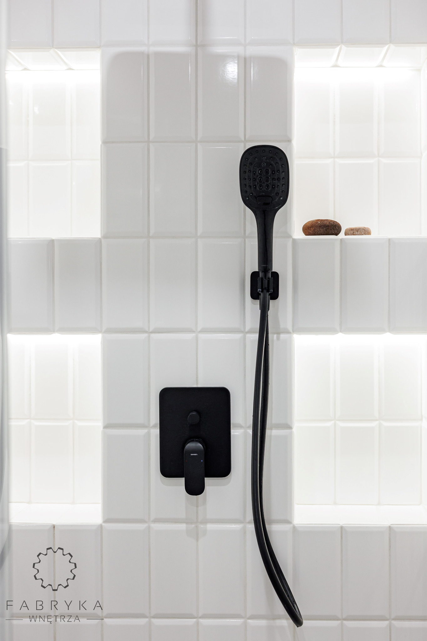 Białe, kafelkowane ściany kabiny prysznicowej z wnęką oświetloną LED-ami, czarna deszczownica z wężem i bateria podtynkowa, dwa kamienie dekoracyjne na półce.