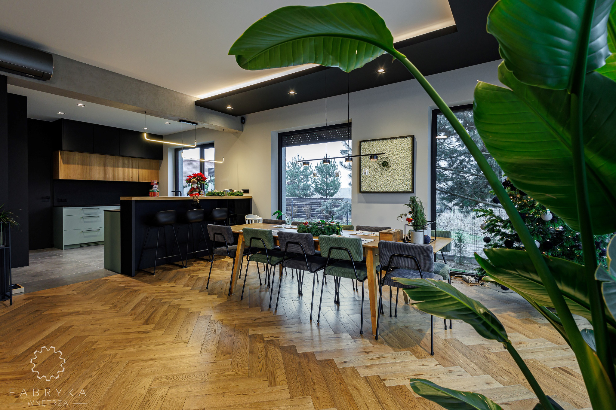 Nowoczesna jadalnia z parkietem w jodełkę, czarną zabudową kuchenną i dużym oknem z widokiem na zaśnieżony ogród, częściowo zasłonięta przez roślinę doniczkową.