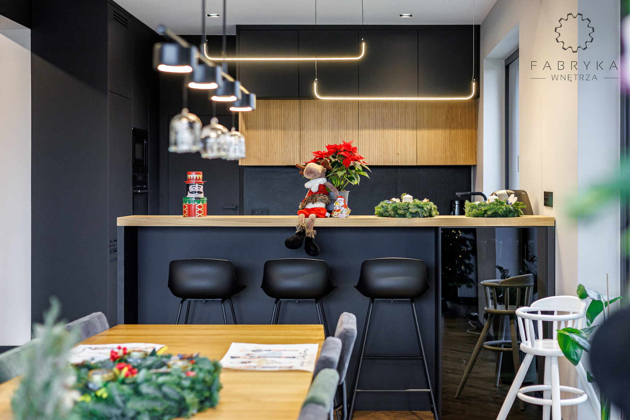 Nowoczesna kuchnia z wyspą, czarnymi szafkami i drewnianymi akcentami oświetlona wiszącymi lampami, udekorowana świątecznymi ozdobami: reniferem, gwiazdą betlejemską i adwentowym wieńcem.
