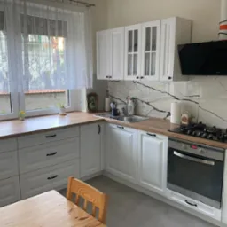 Jasna kuchnia z białymi szafkami, drewnianym blatem i marmurowym backsplashiem, widok na okno z firanką i częściowo widoczne krzesło przy stole.