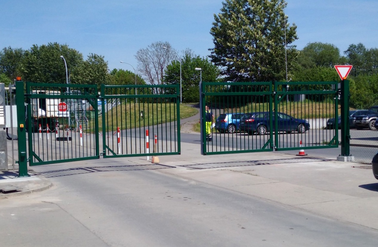 Otwarta, dwuskrzydłowa, metalowa brama wjazdowa w kolorze ciemnej zieleni, z pionowymi prętami, na tle parkingu z samochodami i zieleni. Na bramie widoczny znak STOP.