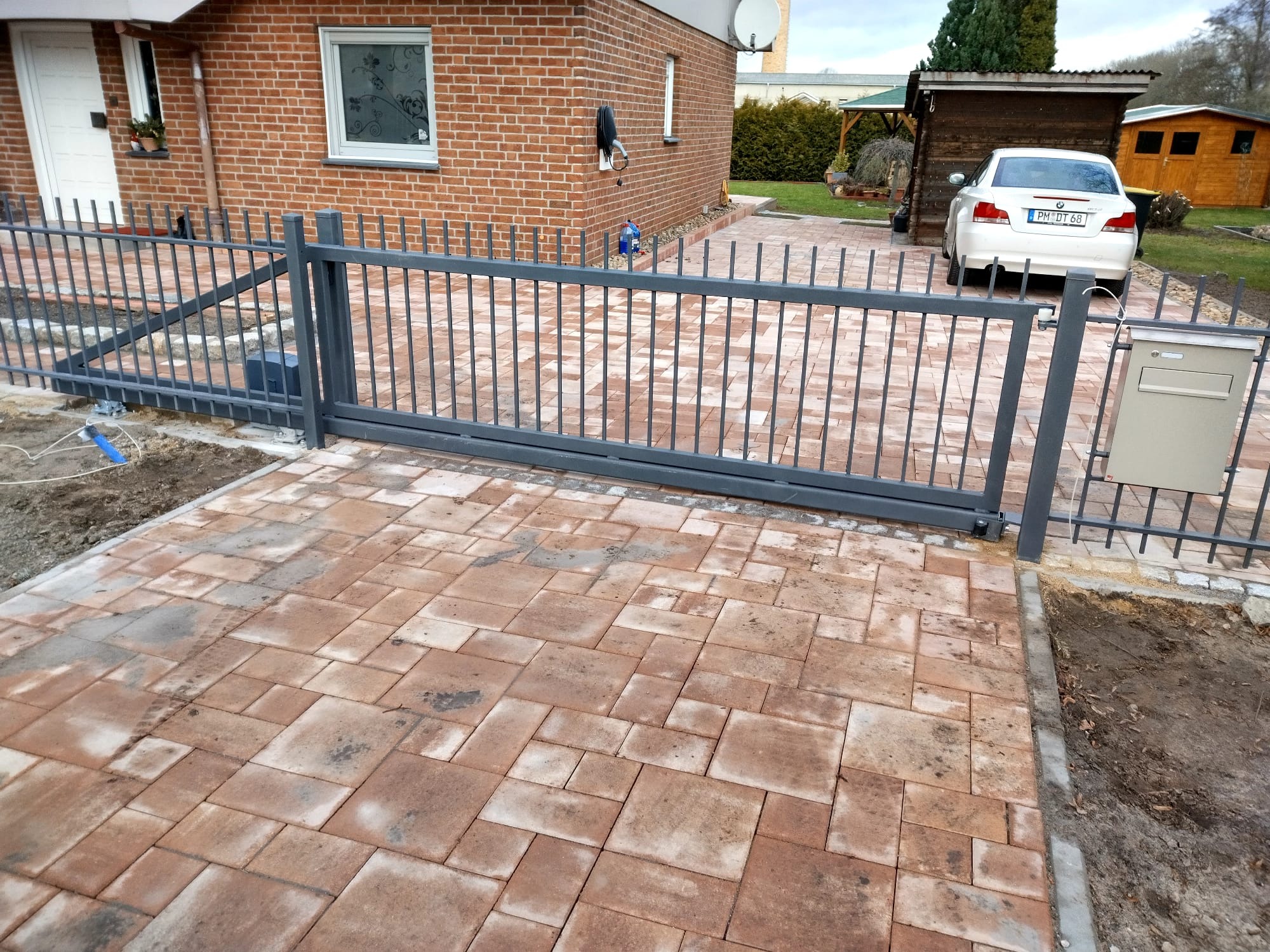 Automatyczna brama przesuwna z szarego metalu przed domem z czerwonej cegły i wybrukowanym podjazdem.