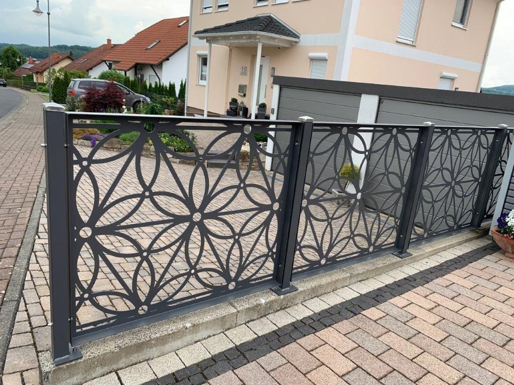 Metalowe ogrodzenie z ażurowym wzorem kwiatowym, pomalowane na szaro, oddzielające posesję od ulicy z kostki brukowej.