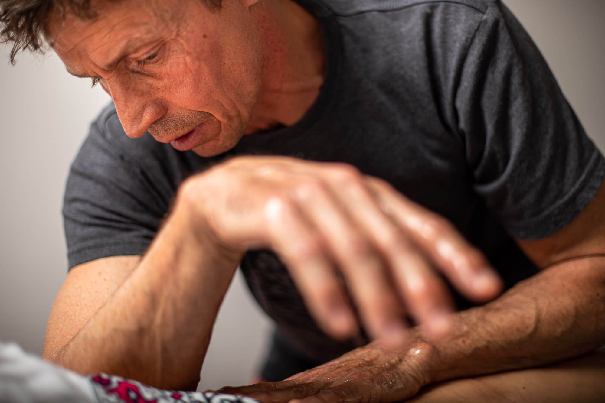 Mężczyzna w szarej koszulce wykonuje ruchy masujące na czyjejś ręce, skupiając się na obszarze nadgarstka i przedramienia, widoczne zmarszczki i żyły na dłoniach.