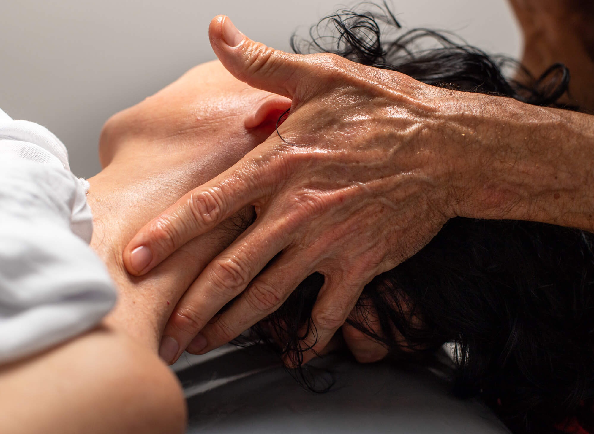 Dłoń terapeuty uciska szyję pacjentki leżącej na plecach, widoczne zmarszczki na dłoni i fragment białej koszuli.