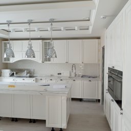 Wykończenia wnętrz Tomasz Michalewski - Jasna kuchnia w stylu klasycznym z białymi szafkami, marmurowym blatem i wiszącymi lampami w kształcie dzwonów, jeszcze częściowo zabezpieczona folią ochronną, widoczne niedoskonałości wykończenia.
