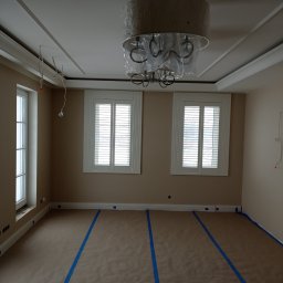 Wykończenia wnętrz Tomasz Michalewski - Pokój przygotowany do malowania: podłoga zabezpieczona brązowym papierem i niebieską taśmą malarską, ściany w odcieniach beżu, białe okna z żaluzjami, dekoracyjny sufit z listwami i wiszący...