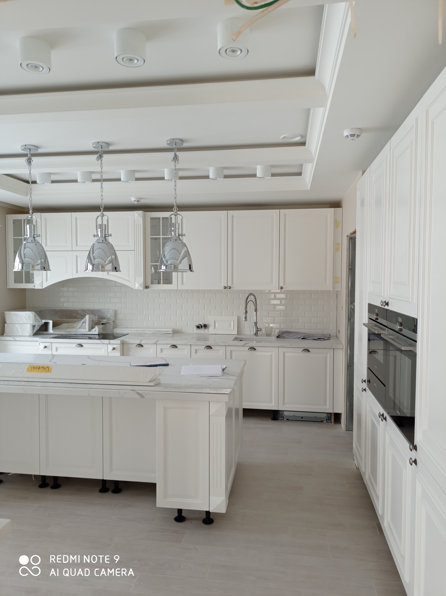 Jasna kuchnia w stylu klasycznym z białymi szafkami, marmurowym blatem i wiszącymi lampami w kształcie dzwonów, jeszcze częściowo zabezpieczona folią ochronną, widoczne niedoskonałości wykończenia.