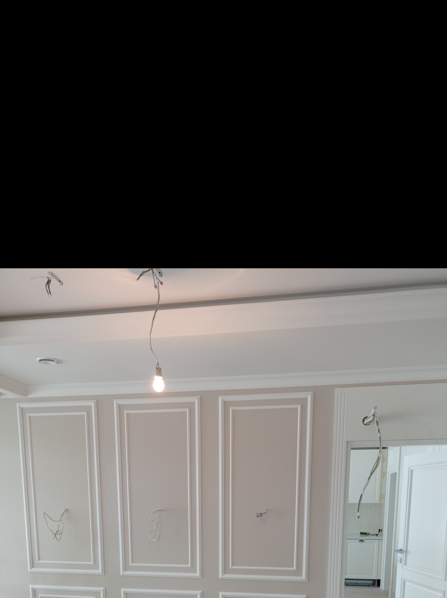 Ściana z dekoracyjnymi listwami w trakcie montażu, widoczne wystające kable i tymczasowe oświetlenie sufitowe, fragment otwartego przejścia do kuchni w tle.