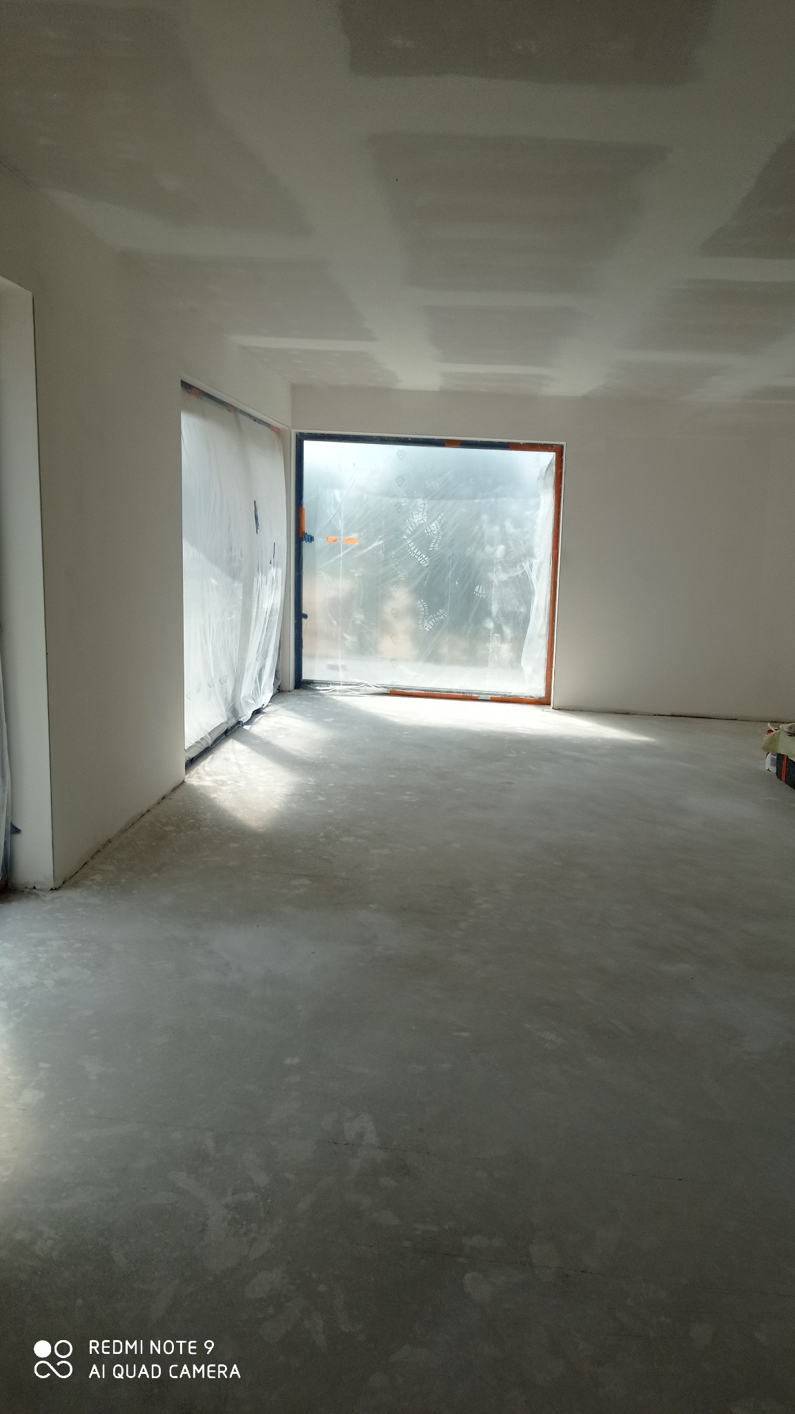 Pokój w trakcie prac wykończeniowych: świeżo wylana wylewka na podłodze, ściany i sufit po gładziowaniu, okno zabezpieczone folią malarską.