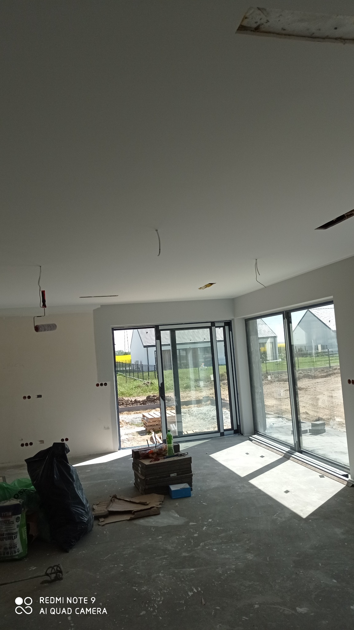 Wnętrze w trakcie prac wykończeniowych: surowa podłoga, świeżo pomalowane ściany i sufit z widocznymi punktami elektrycznymi, duże okna przesuwne z widokiem na ogród, narzędzia i materiały...