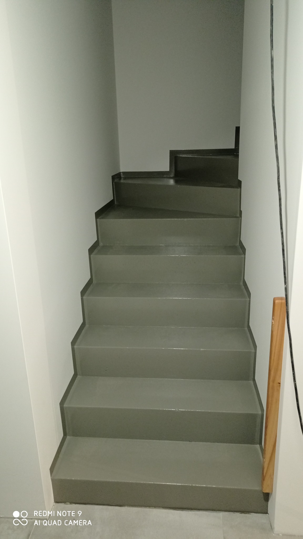 Schody wewnętrzne pomalowane na szaro, widoczne z dołu, z fragmentem drewnianej barierki po prawej stronie.