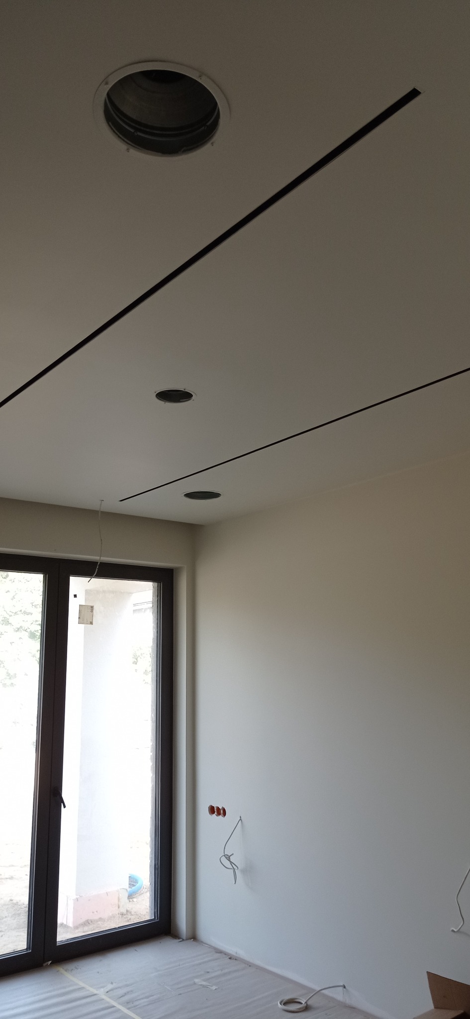 Biały sufit z trzema okrągłymi otworami na oświetlenie i czarnymi, wąskimi listwami dekoracyjnymi; widoczne kable elektryczne wystające ze ściany oraz zabezpieczona folią podłoga w trakcie prac...