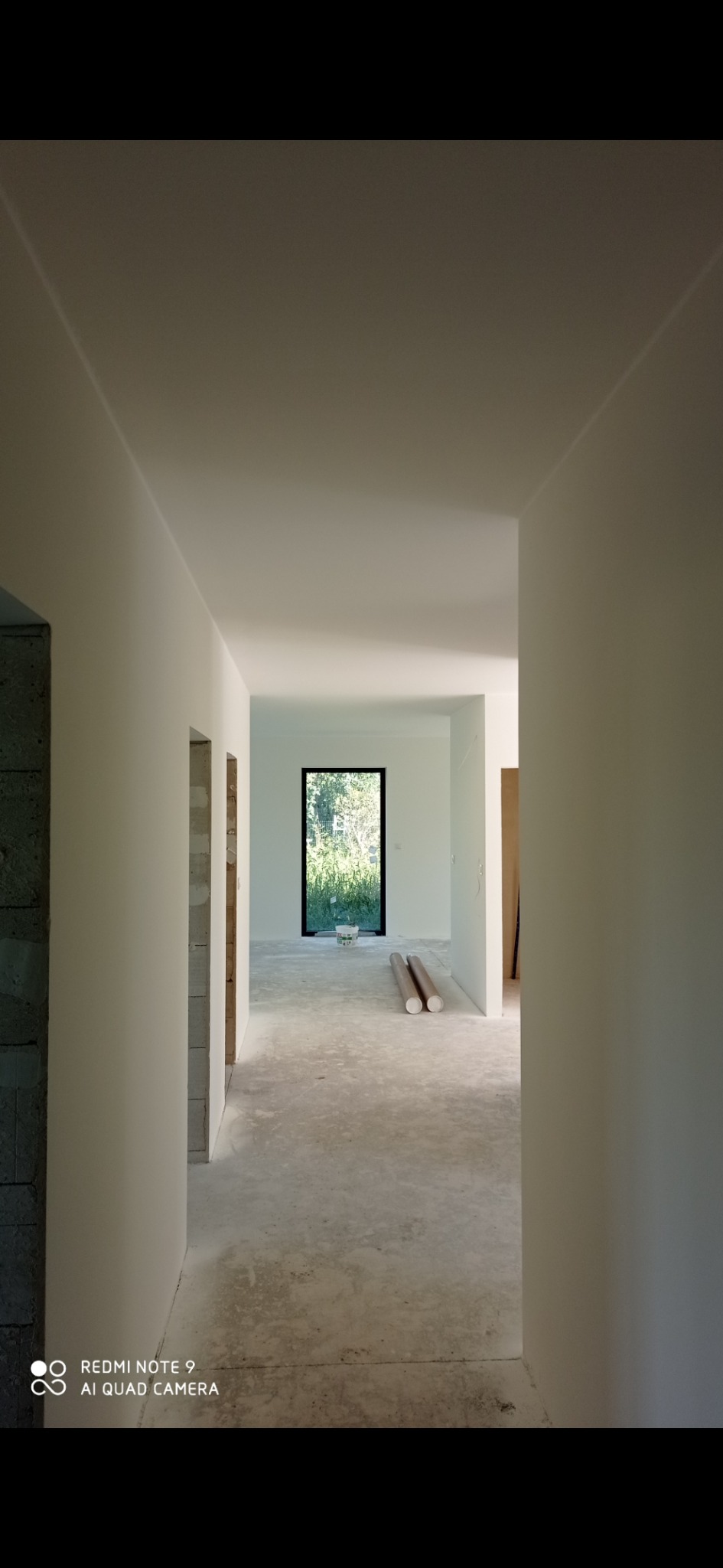 Wnętrze w trakcie prac wykończeniowych: świeżo pomalowane ściany na biało, surowy beton na podłodze, widoczne otwory drzwiowe i okno z widokiem na zieleń, dwa zwinięte rulony materiału leżące...