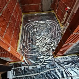 Janosz - Instalacja ogrzewania podłogowego w surowym pomieszczeniu z czerwonej cegły, widoczne rury grzewcze na folii izolacyjnej i siatce montażowej, częściowo zakryte przez stopę osoby.