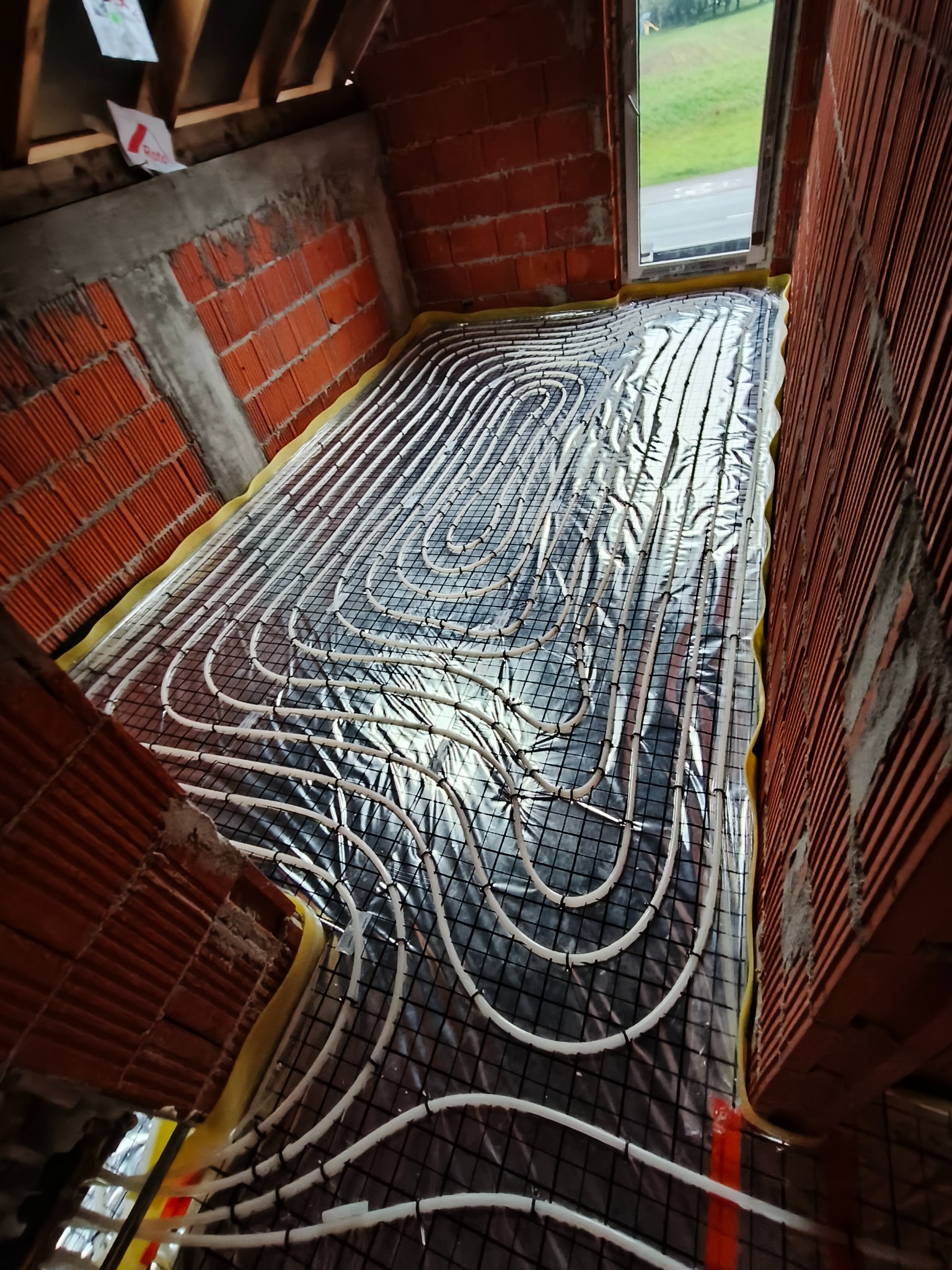 Montaż rur grzewczych podłogowych na folii izolacyjnej w budynku w Krowiarkach, widoczne czerwone cegły ścian i okno w tle.