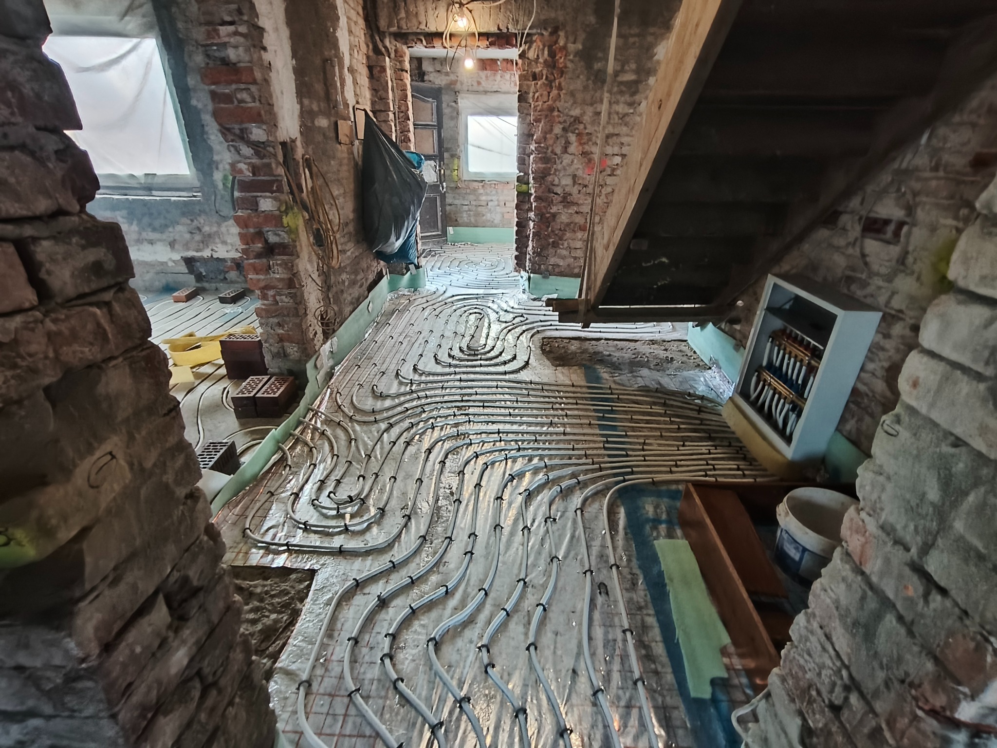 Instalacja ogrzewania podłogowego w trakcie remontu, widoczne białe rury na folii izolacyjnej, surowe cegły ścian, w tle okno i elementy instalacji grzewczej.