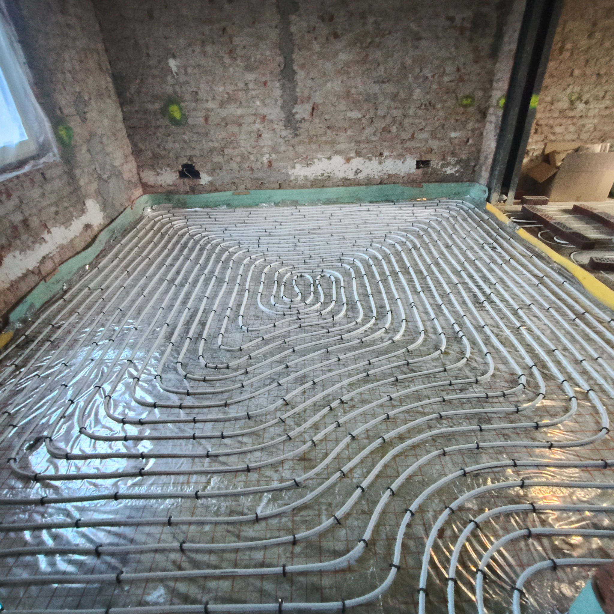 Ułożone rury ogrzewania podłogowego na folii, przymocowane czarnymi uchwytami, w surowym wnętrzu z ceglaną ścianą, gotowe do zalania posadzką.